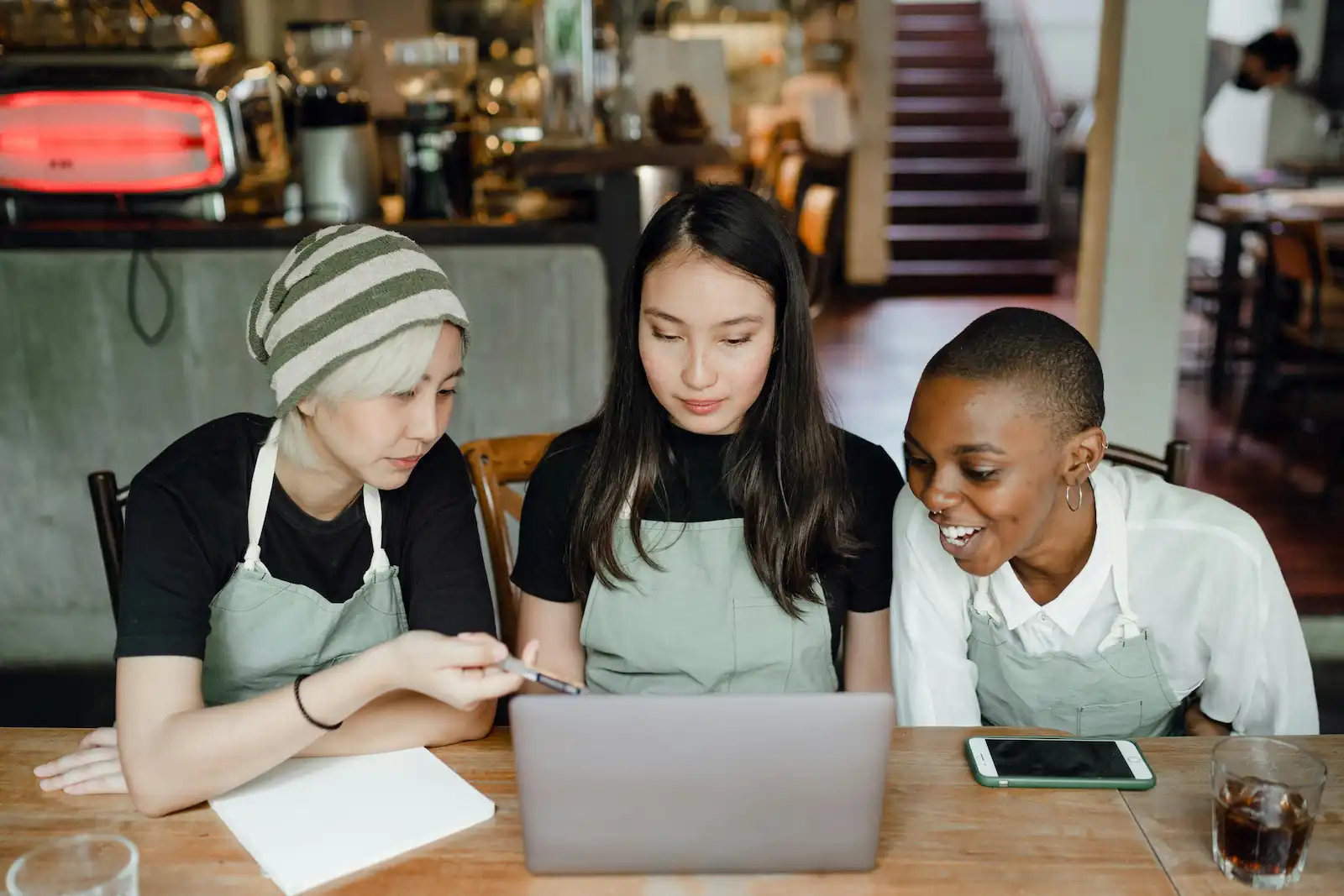 3 small business employees are sitting in a cafe, engaging with the same laptop screen.