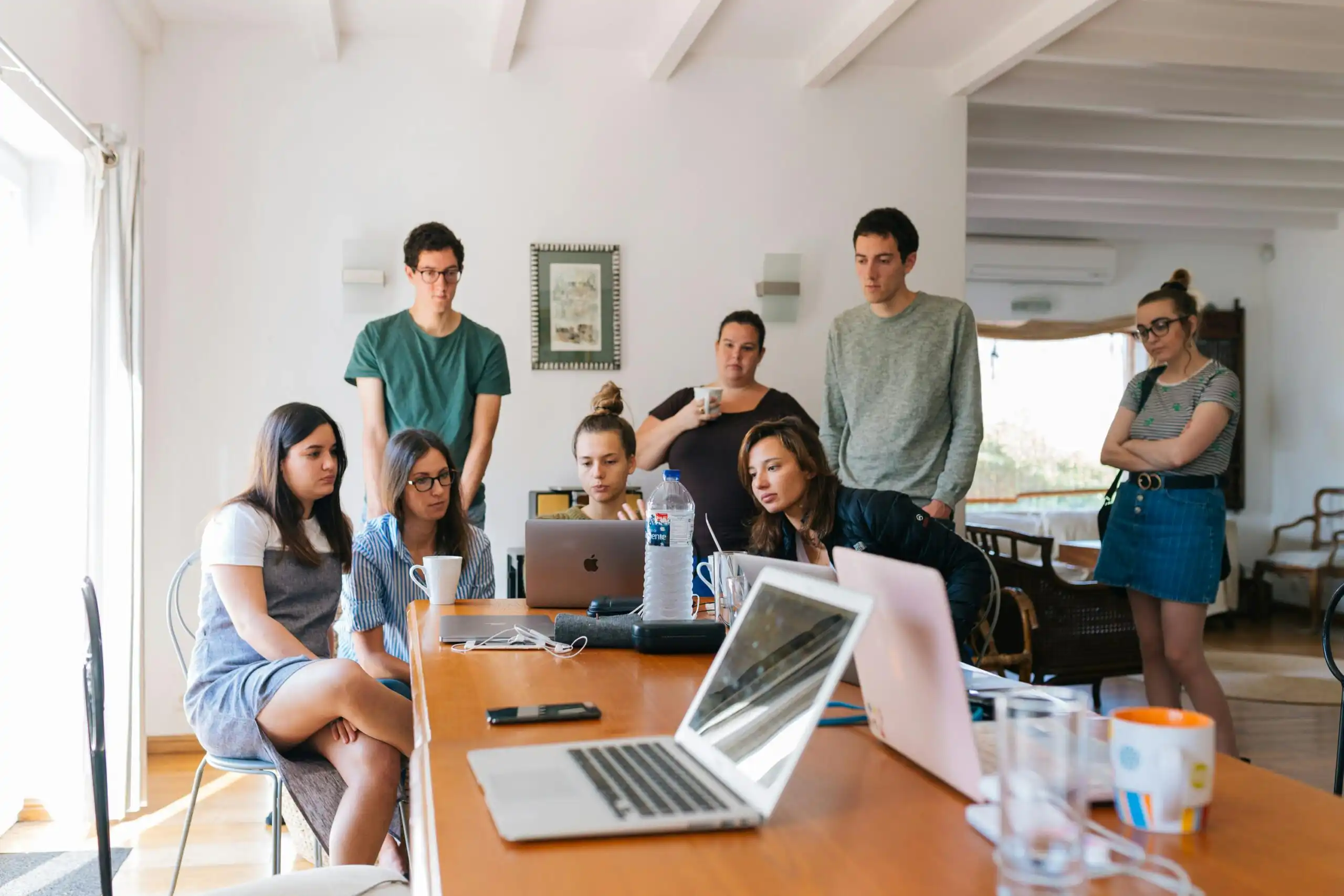 A team of 8 marketing professionals are sitting in a meeting. They are looking towards the same laptop screen discussing their company website.