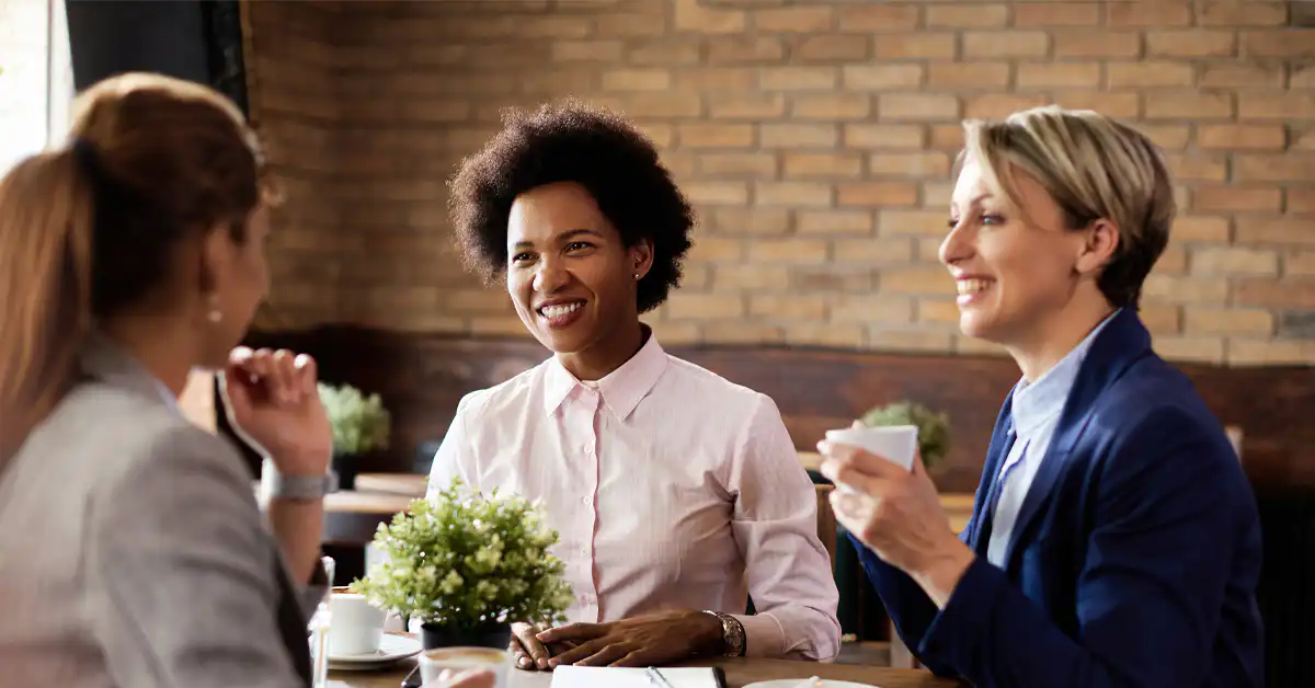 A photo of three people with different hair styles in a café, including a woman with curly, short, black hair, who are having a nice, bright chat.