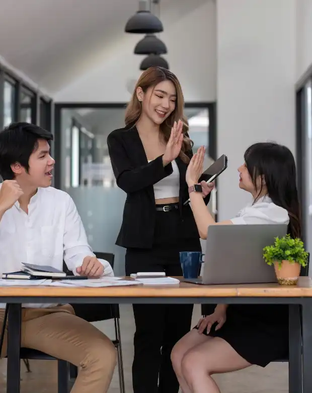 Three young Asian business professionals smiling and giving a high-five during a team meeting with a laptop, representing Traditional Chinese translation and business services